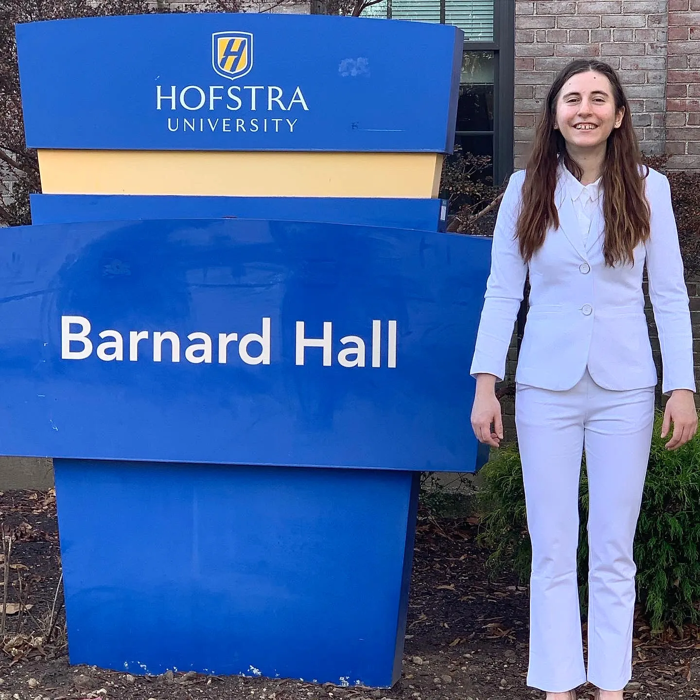 Lillian Riggs in a white suit beside the Hofstra University Barnard Hall sign